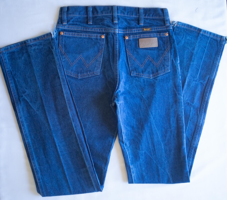 May include: A pair of blue denim jeans featuring a classic five-pocket design with copper-coloured rivets. The back pockets display a signature W-shaped stitching pattern in orange thread, and a brown leather-like patch with the Wrangler logo.