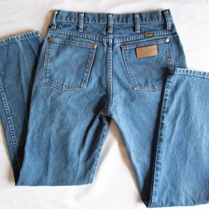 Puede incluir: Vaqueros de mezclilla azul con cintura alta, trabillas para cintur&oacute;n y dos bolsillos traseros con remaches de color cobre. El bolsillo trasero derecho tiene un parche de cuero marr&oacute;n con el logo Wrangler y ambos bolsillos tienen una costura en W.