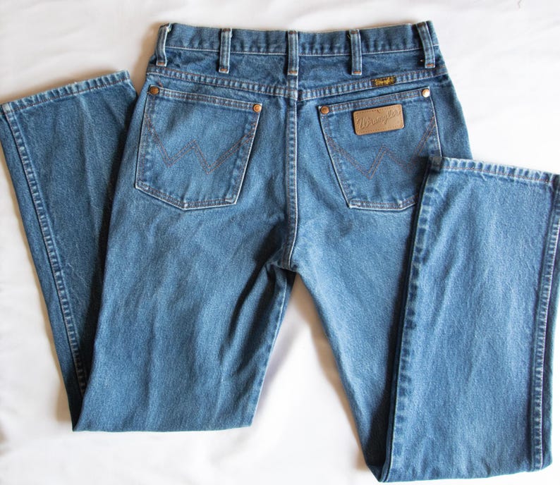 Puede incluir: Vaqueros de mezclilla azul con cintura alta, trabillas para cintur&oacute;n y dos bolsillos traseros con remaches de color cobre. El bolsillo trasero derecho tiene un parche de cuero marr&oacute;n con el logo Wrangler y ambos bolsillos tienen una costura en W.