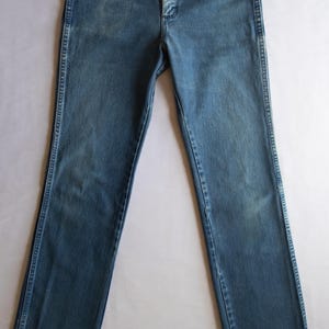 Puede incluir: Unos vaqueros de mezclilla azul con dise&ntilde;o de cintura alta, cierre de botones, remaches de color cobre en los bolsillos delanteros y corte de pierna recta. La tela presenta un lavado medio con un ligero desgaste en los muslos.