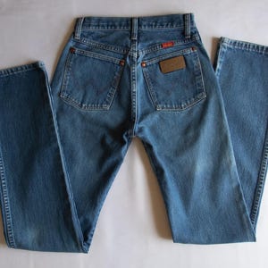 May include: A pair of blue denim jeans featuring a high-waisted design, two back pockets with copper-coloured rivets, and a brown leather patch on the right pocket with the Wrangler brand name clearly visible in embossed lettering.