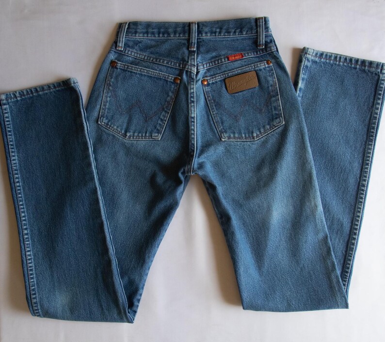 May include: A pair of blue denim jeans featuring a high-waisted design, two back pockets with copper-coloured rivets, and a brown leather patch on the right pocket with the Wrangler brand name clearly visible in embossed lettering.