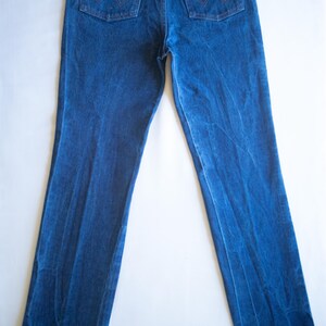 May include: A pair of dark blue denim jeans featuring a high-rise waist, straight legs, and two back pockets with copper-coloured rivets. The right back pocket displays a brown leather patch with the Wrangler brand name and a stitched W design.