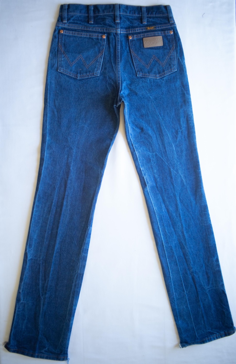 May include: A pair of dark blue denim jeans featuring a high-rise waist, straight legs, and two back pockets with copper-coloured rivets. The right back pocket displays a brown leather patch with the Wrangler brand name and a stitched W design.