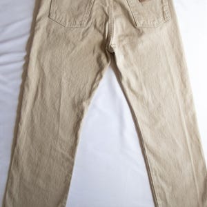 Op de afbeelding: Een beige spijkerbroek van denim met een klassiek ontwerp met vijf zakken, een knoopsluiting en riemlussen. De achterkant toont twee achterzakken met koperkleurige klinknagels en een beige leren label op de rechterzak met het Wrangler-merklogo.