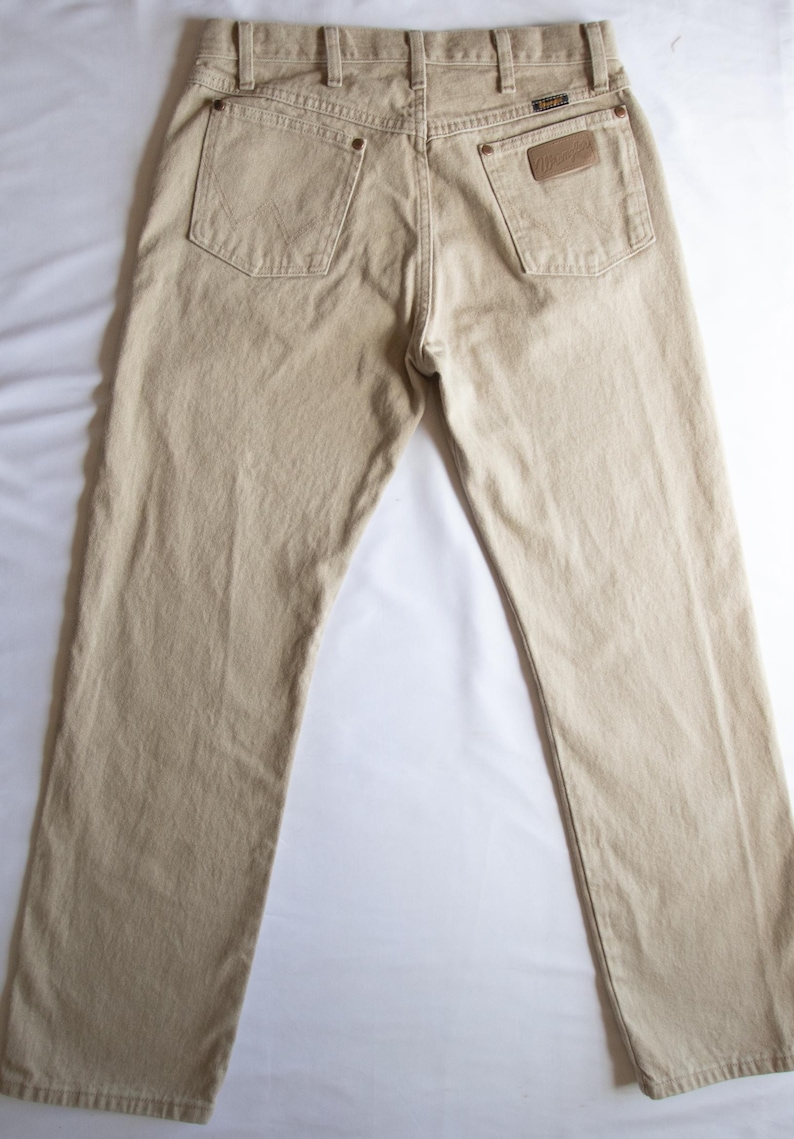 Op de afbeelding: Een beige spijkerbroek van denim met een klassiek ontwerp met vijf zakken, een knoopsluiting en riemlussen. De achterkant toont twee achterzakken met koperkleurige klinknagels en een beige leren label op de rechterzak met het Wrangler-merklogo.