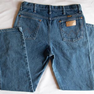Pu&ograve; includere: Un paio di jeans in denim blu di media tonalit&agrave; con un classico design a cinque tasche, passanti per cintura e rivetti color rame. La tasca posteriore destra presenta una toppa in pelle beige con il marchio Wrangler e una cucitura a W.