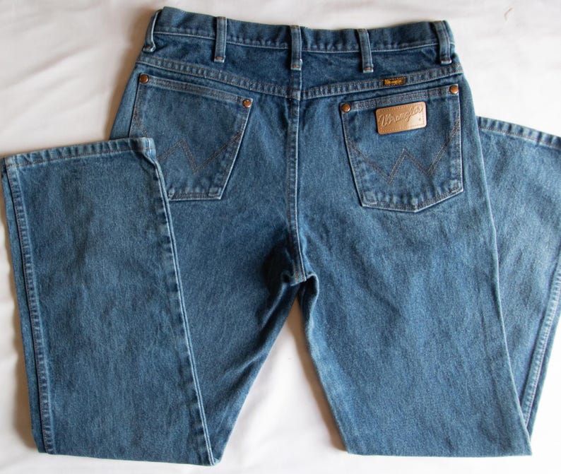 Pu&ograve; includere: Un paio di jeans in denim blu di media tonalit&agrave; con un classico design a cinque tasche, passanti per cintura e rivetti color rame. La tasca posteriore destra presenta una toppa in pelle beige con il marchio Wrangler e una cucitura a W.