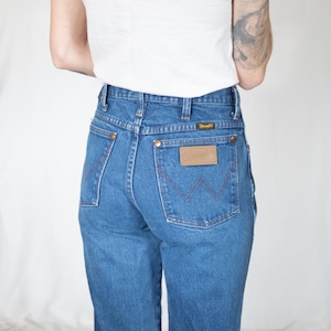 Puede incluir: Vaqueros de mezclilla azul de talle alto con un dise&ntilde;o cl&aacute;sico de cinco bolsillos, remaches de color cobre y un parche de cuero marr&oacute;n en el bolsillo trasero con el nombre de la marca Wrangler. Los bolsillos traseros tienen bordado en forma de W.