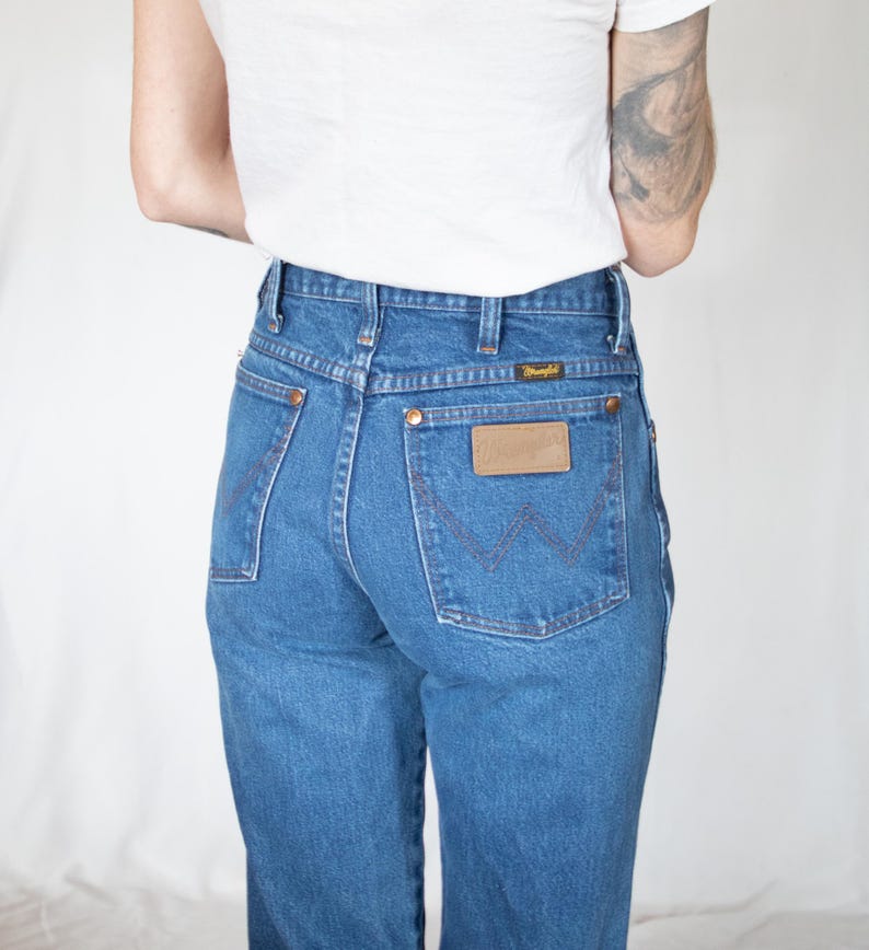 Puede incluir: Vaqueros de mezclilla azul de talle alto con un dise&ntilde;o cl&aacute;sico de cinco bolsillos, remaches de color cobre y un parche de cuero marr&oacute;n en el bolsillo trasero con el nombre de la marca Wrangler. Los bolsillos traseros tienen bordado en forma de W.