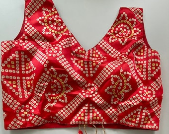 May include: A sleeveless red blouse with a V-neck and a repeating white and yellow geometric pattern. The blouse has decorative tassels hanging from the bottom. The tassels are red with a white and yellow pattern, and gold-colored metal accents.