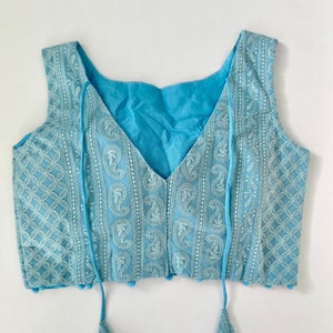 Pu&ograve; includere: Un top senza maniche azzurro con ricami bianchi intricati. Il top presenta uno scollo a V e nappe decorative. Il tessuto presenta un motivo paisley e geometrico. &Egrave; un capo leggero e arioso.