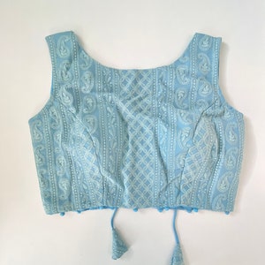 Pu&ograve; includere: Un top senza maniche azzurro con ricami bianchi intricati. Il design presenta motivi paisley e geometrici sul tessuto. Il top ha uno scollo tondo e nappe decorative sull'orlo.