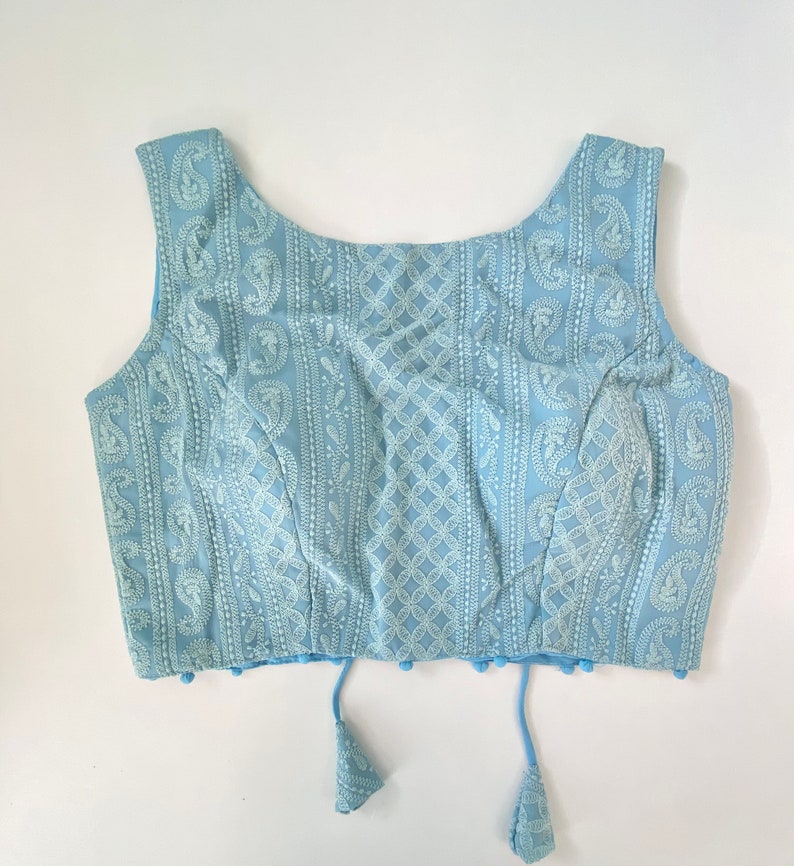 Pu&ograve; includere: Un top senza maniche azzurro con ricami bianchi intricati. Il design presenta motivi paisley e geometrici sul tessuto. Il top ha uno scollo tondo e nappe decorative sull'orlo.