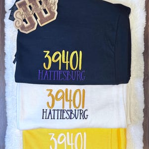 May include: Three folded t-shirts in black, white, and yellow. Each shirt features embroidered text reading 39401 and HATTIESBURG in contrasting colors. A wooden tag with the text Stitched by JB is placed on the top black t-shirt.