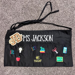 May include: Black teacher's apron with multiple pockets and a black tie. The apron is embroidered with the name "MS. JACKSON" in white, along with various school-themed patches, including "ABC", a lightbulb, an apple, and "#1 teacher".