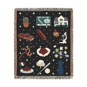 May include: A black woven tapestry with a white fringe border. The tapestry features a variety of colorful illustrations, including a cactus, a house, a truck, a ketchup bottle, an onion, a microscope, a stick figure, a hand holding a heart, a bouquet of flowers, a plate of spaghetti, a lightning bolt, a baseball bat and ball, and a dress. The text "The City of Forks Welcomes You" is also included.