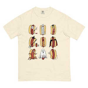 May include: Cream-colored t-shirt featuring a grid of Halloween-themed hot dogs. Each hot dog is dressed as a different spooky character, including a mummy, vampire, and ghost. The design is playful and colorful, perfect for Halloween.