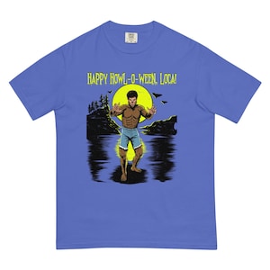 May include: A periwinkle blue t-shirt featuring a graphic of a werewolf standing in water before a large yellow moon. The text "HAPPY HOWL-O-WEEN, LOCA!" is printed in yellow above the figure, which is depicted with brown fur and blue shorts.