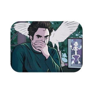May include: A rectangular bath mat featuring a comic-style illustration. The artwork depicts a person covering their mouth, with a white bird and a framed skeleton drawing in the background. The color palette includes shades of green, white, and black.