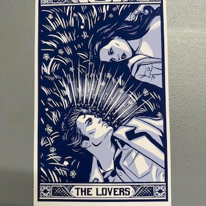 May include: A blue and white tarot card print featuring two figures lying in a field of grass and flowers. The card is labeled "VI" at the top and "THE LOVERS" at the bottom.
