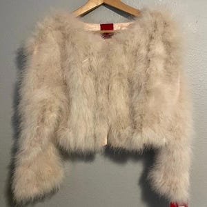 May include: A cropped jacket covered in long, off-white feathers with a round neckline and long sleeves. The garment is displayed on a wooden hanger and features a small red tag attached to the lower right side and a red label at the inner neck.