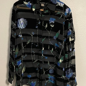 Puede incluir: Camisa negra con botones y estampado floral. La camisa presenta rayas horizontales negras y diseños florales azules. La camisa de manga larga está colgada en una percha de madera.