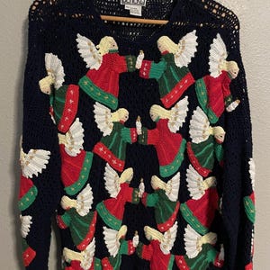 Könnte beinhalten: Marineblauer Häkelpullover mit mehreren Engelsfiguren in Rot, Grün und Weiß. Die Engel halten Kerzen und haben weiße Flügel. Der Pullover hat einen gewellten Saum und ein lockeres, offenes Webdesign.