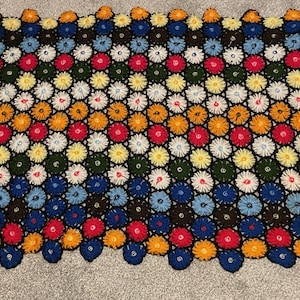 Puede incluir: Una colorida manta de ganchillo hecha a mano con un patrón floral repetido. La manta muestra una variedad de colores, incluyendo rojo, amarillo, azul, verde y naranja, sobre un fondo negro. Los bordes son festoneados, añadiendo un toque decorativo.