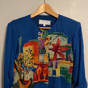 May include: A blue long-sleeve jacket with a colorful abstract print. The print features a floral and architectural design in shades of red, orange, green, and yellow. The jacket has a v-neck and is labeled "Platinum".