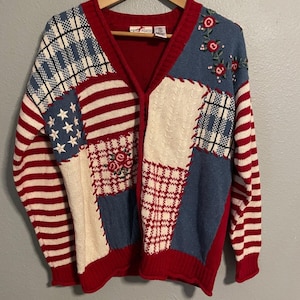 Può includere: Un cardigan rosso, bianco e blu con un design patchwork. La maglia presenta strisce, stelle e ricami floreali. Il cardigan con scollo a V è appeso a una gruccia di legno.