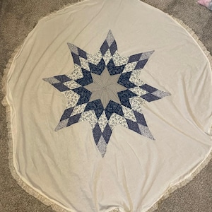 May include: A round, off-white tree skirt with a large, eight-pointed star applique. The star is made of various blue and white patterned fabrics. The edge of the skirt is trimmed with a delicate lace.