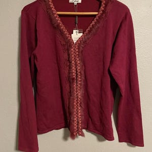 Può includere: Un cardigan bordeaux a maniche lunghe con scollo a V e finiture decorative. La finitura presenta una combinazione di pizzo e una texture piumata. Il cardigan è appeso a una gruccia di legno.