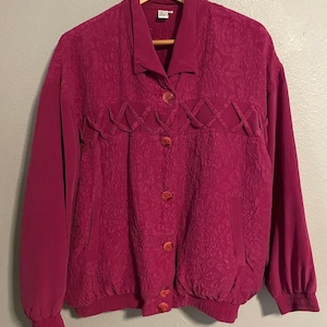 May include: A magenta long-sleeve jacket featuring a textured floral pattern on the front panels, a collared neckline, and a button-down front closure. The garment includes a decorative lattice detail across the chest and elasticized cuffs.