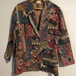 May include: A long-sleeve blazer featuring a multi-colored tapestry pattern in shades of red, blue, green, and gold. The garment has a wide lapel, two silver-toned metal buttons for closure, and two front pockets, all hanging on a wooden hanger.