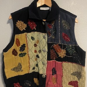 Puede incluir: Un chaleco patchwork con cuello y frente negros, con cuadrados texturizados en amarillo, rosa, verde y beige. El chaleco está adornado con diseños de hojas bordadas en colores otoñales y tiene cierres de botones.