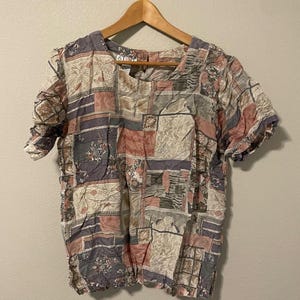 May include: A short-sleeved blouse featuring a multi-colored patchwork pattern in shades of dusty rose, muted purple, beige, and dark gray. The garment has a round neckline, a button closure at the back of the neck, and a gathered hemline.