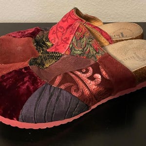 Op de afbeelding: Een paar patchwork klompen in tinten rood, bordeaux en donkerblauw. De klompen hebben een kurken voetbed en een roze rubberen zool. De bovenkant is gemaakt van verschillende stoffen, wat een unieke en getextureerde uitstraling creëert.