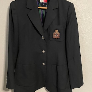 May include: A black blazer with a classic design, featuring a notched lapel, two front pockets, and three silver buttons. The blazer has a crest emblem on the chest pocket and a Tommy Hilfiger label.