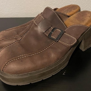 Puede incluir: Zuecos de cuero marrón con tacón grueso y cierre de hebilla. Los zuecos tienen punta redondeada y detalles de costura blancos. Las suelas son de color marrón oscuro y los tacones miden unos 7,6 cm de alto.