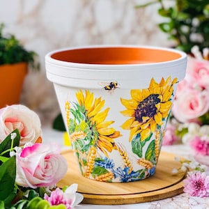 May include: White ceramic plant pot decorated with sunflowers, wheat, and blue wildflowers. A bumblebee flies near the rim. The pot is on a wooden tray, surrounded by flowers. The pot's interior is orange.