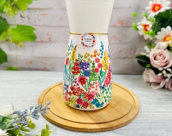 Op de afbeelding: Een witte keramische vaas met een kleurrijke botanische print van wilde bloemen in tinten rood, blauw, geel en groen. Een kleine ronde sticker met de tekst "Handmade with love" is met een dun gouden koordje aan de bovenkant bevestigd.
