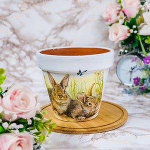 May include: White painted flower pot with a detailed illustration of two brown rabbits in a field. The pot has a light brown interior and sits on a wooden coaster. A small butterfly is also depicted on the pot.