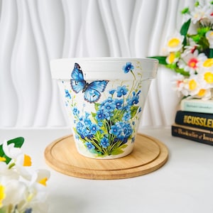 May include: A white flower pot featuring a vibrant blue butterfly and clusters of small blue flowers with green leaves. The pot has a wide rim and a tapered body, decorated with a detailed botanical print on its exterior surface.