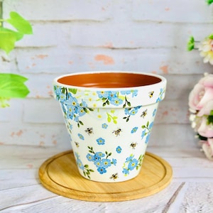 May include: A white ceramic plant pot adorned with blue forget-me-not flowers and bee illustrations. The pot rests on a round wooden coaster. The interior of the pot is brown.