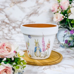 May include: White ceramic plant pot decorated with a Peter Rabbit design, surrounded by colorful flowers and a butterfly. The pot rests on a round wooden coaster. The inside of the pot is brown.