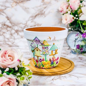 May include: A white ceramic plant pot decorated with a vibrant birdhouse design. The pot displays illustrations of birdhouses, flowers, and birds. It rests on a round wooden coaster, with flowers in the background.