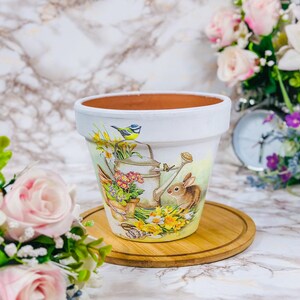 May include: White ceramic plant pot featuring a painted spring garden scene. The design includes a watering can, bunny, bird, and flowers. The pot sits on a round wooden base, suitable for indoor or outdoor use.