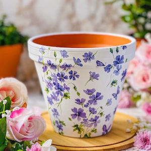 May include: A white ceramic plant pot with a bright orange interior. The exterior is decorated with delicate purple floral patterns. The pot is designed for planting and adds a touch of elegance to any garden or indoor space.