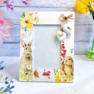 May include: White wooden picture frame with a floral and rabbit design. The frame features watercolour illustrations of rabbits, flowers, and butterflies. Two heart-shaped ornaments hang from a twine bow.